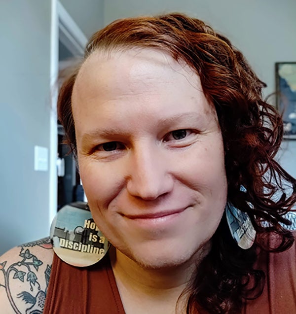 Em Rabelais, a white person, faces the camera in a room painted light grey. They have long, curling orange-red just longer-than-shoulder-length hair on their left side. The hair on their right side is short. The person is smiling, wearing large round earrings with a multicolored background and white bold text stating 