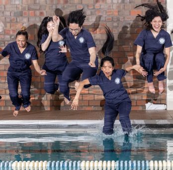 Students wearing dark blue nursing scrubs leap from a pool deck into the pool water 