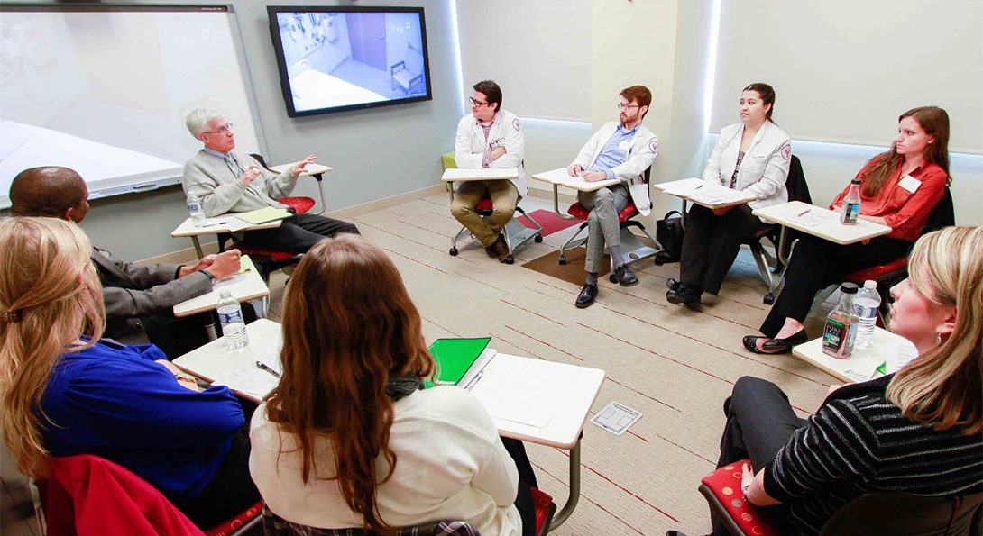 Students sitting at desks arranged in a circle, listening to an instructor, also seated in a desk