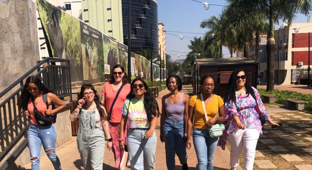 Seven women walking together towards the viewer with buildings and palm trees in the background.