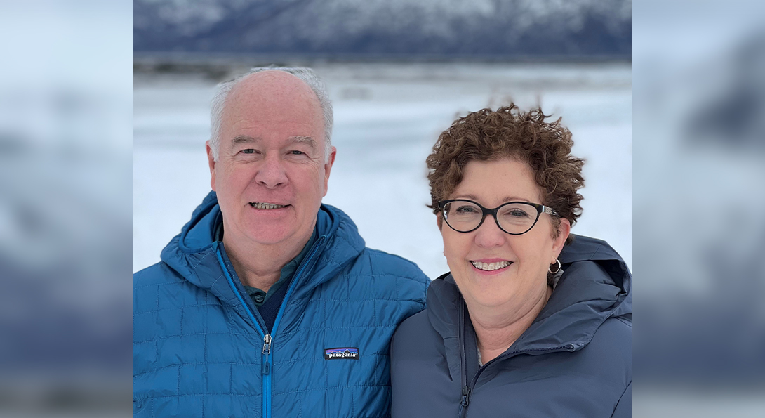 Couple in blue winter jackets smile in front of snowy lake and mountain