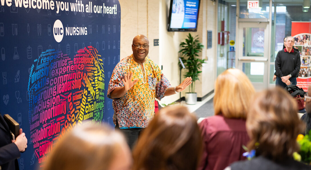 A smiling Black man in yellow, white and blue patterned shirt addresses a blurry crowd. He stands in front of large banner branded with the UIC College of Nursing logo and featuring a heart-shaped word cloud.