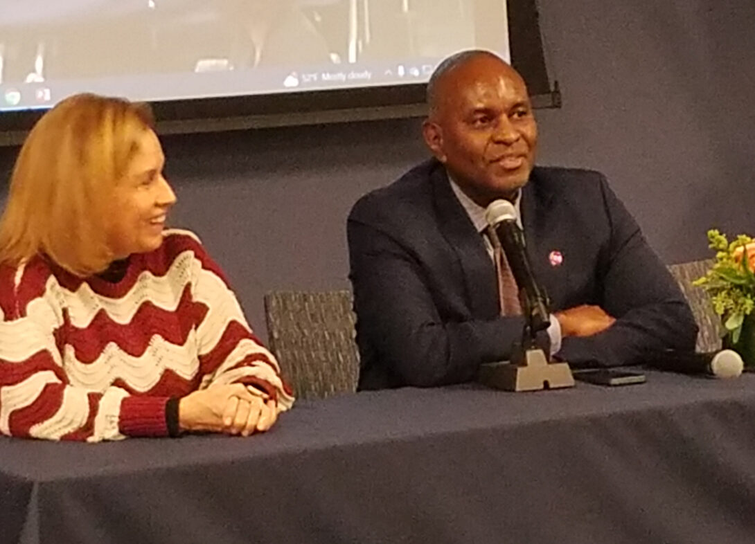 A Black man in a dark suit jacket sits at a table behind a microphone. A blond woman in a red-and-white-striped sweater sits to his right, looking on.