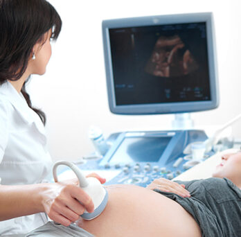 An ultrasound technician administers an ultrasound on the pregnant belly of a woman with blond hair, both watch the image on screen
                  