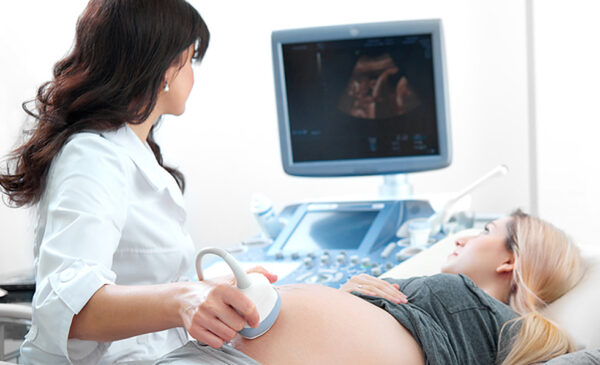 An ultrasound technician administers an ultrasound on the pregnant belly of a woman with blond hair, both watch the image on screen