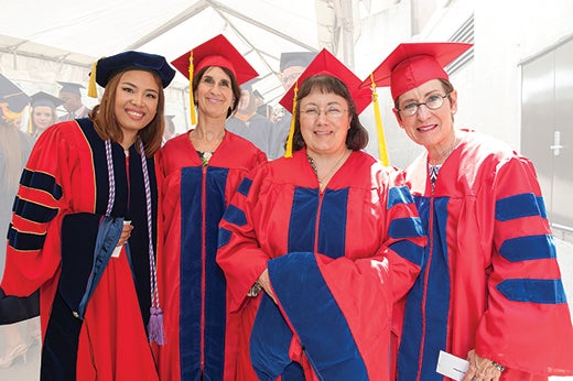 Four women wearing red and blue or red and black graduation gowns and caps pose and smile to the camera