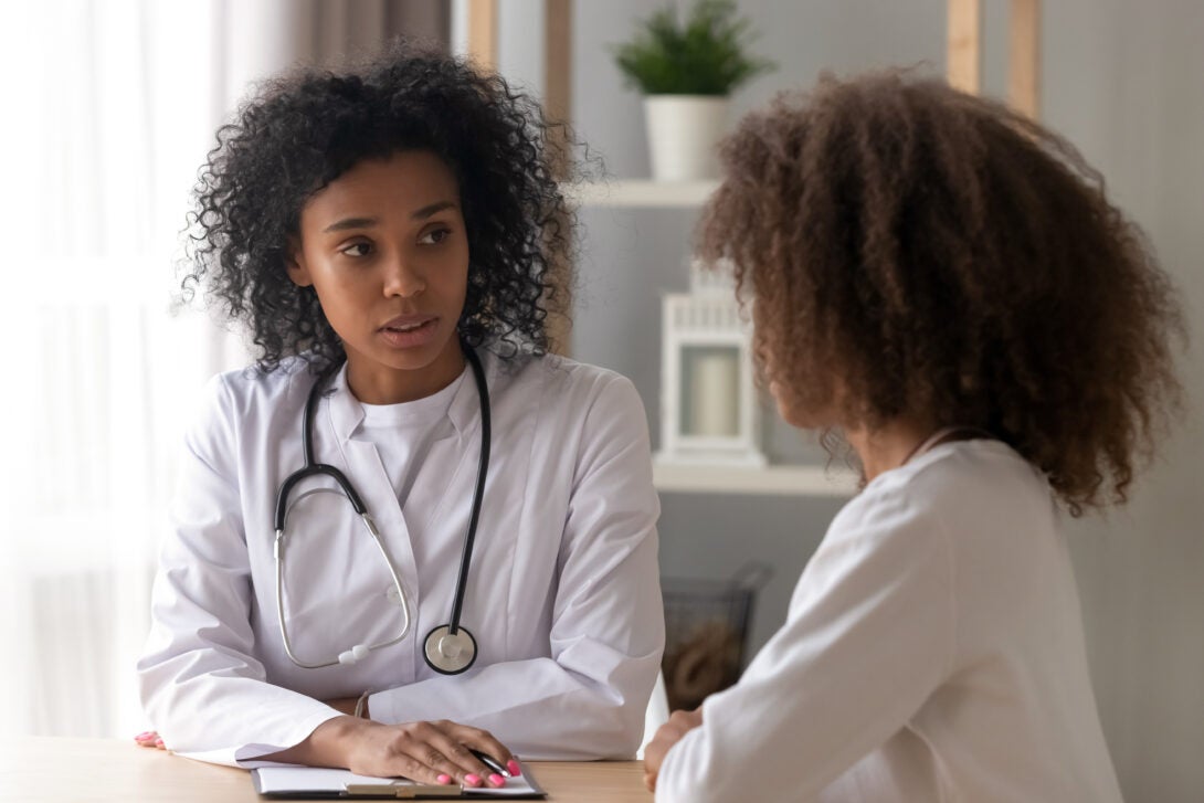 A Black woman in a white coat wearing a stethoscope sits with a clipboard in front of her talking to a younger girl.