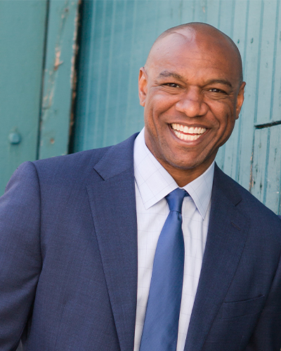 A Black man who is bald and wearing a blue suit and tie smiles at the camera