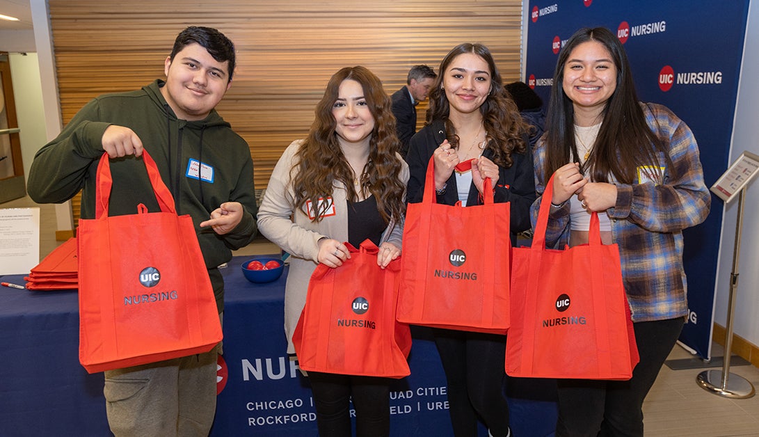 A man and three women, all college-age, pose and smile for the camera holding up red tote bags that say 