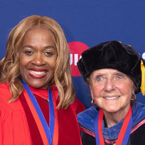 A Black woman with blond hair stands to the right of a shorter white woman, both of them wearing academic robes and medals investiture around their necks