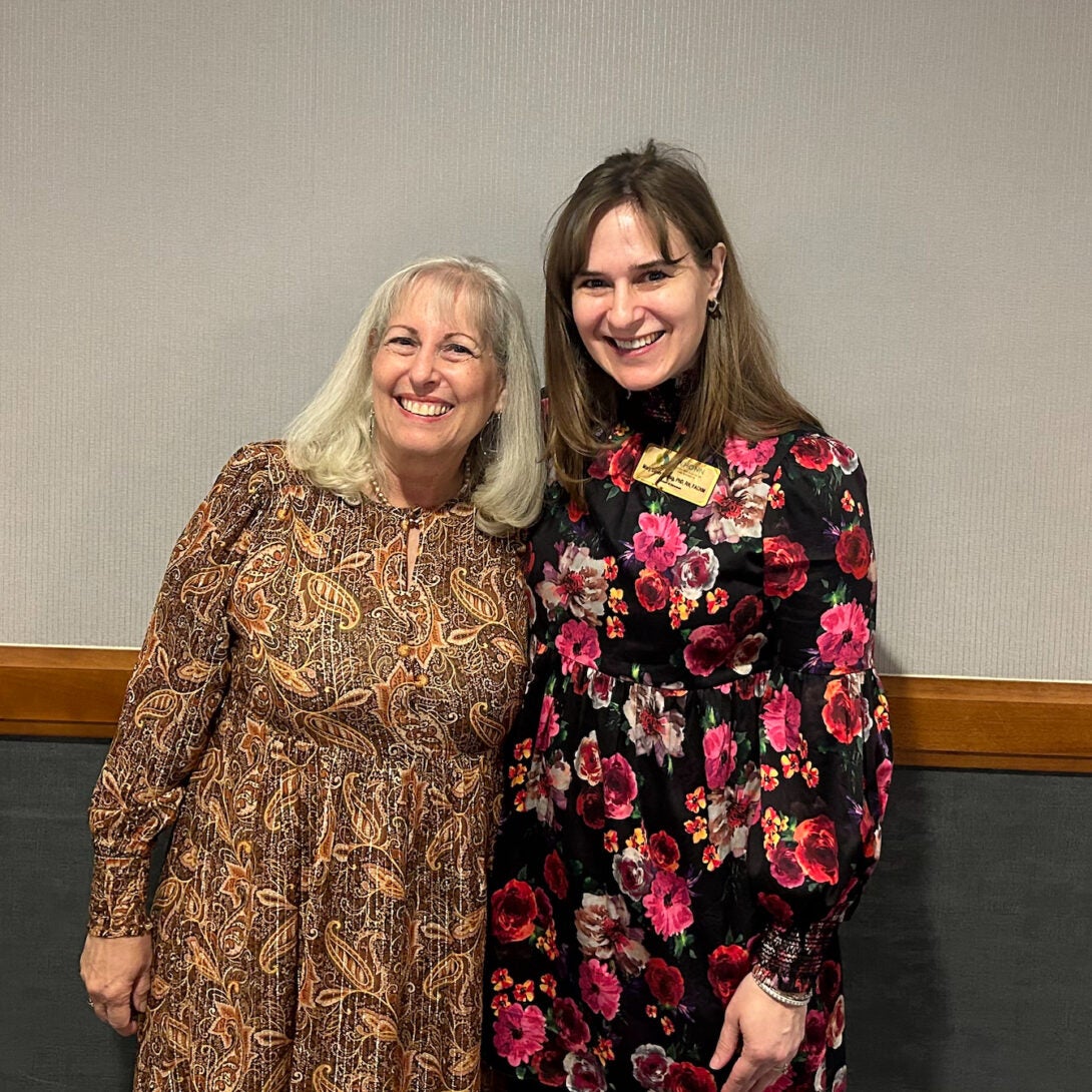 Two women in floral dresses smile with their arms around each other