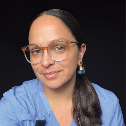 A close up portrait of a female wearing large eyeglasses with her black hair pulled back into a ponytail that wraps around to rest on her shoulder