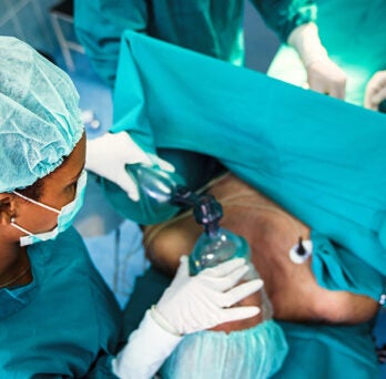 Aerial image of anesthetist wearing blue scrubs, holding an anesthesia mask over the mouth and nose of a surgical patient
                  