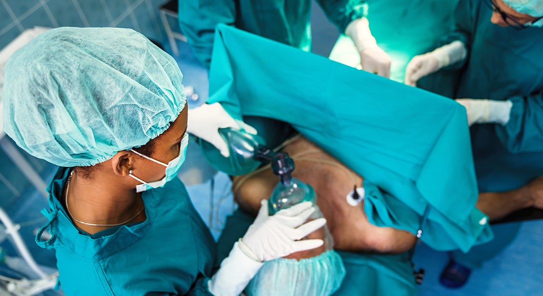 Aerial image of anesthetist wearing blue scrubs, holding an anesthesia mask over the mouth and nose of a surgical patient