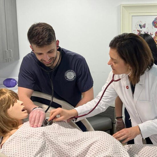 A student in nursing scrubs and an instructor in a white coat smile as they both place stethescopes on the chest of a manikin