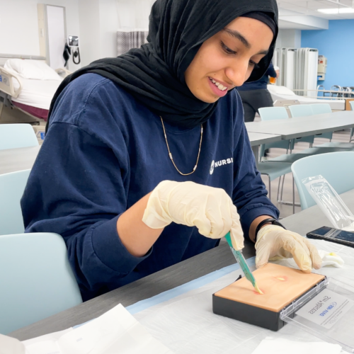 A student wearing UIC Nursing scrubs, a hijab and rubber gloves simulates lancing an absess