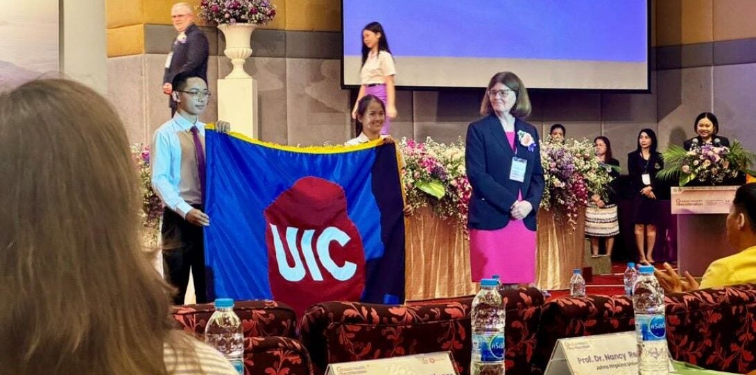 Woman in suit jacket stands next to a flag with the UIC logo