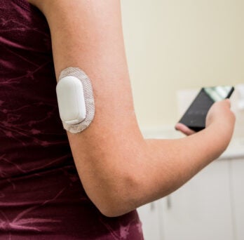 close up of a white woman wearing a sleeveless top, with a white, oval attachment on her exposed upper arm and a smartphone in the hand of the same arm 