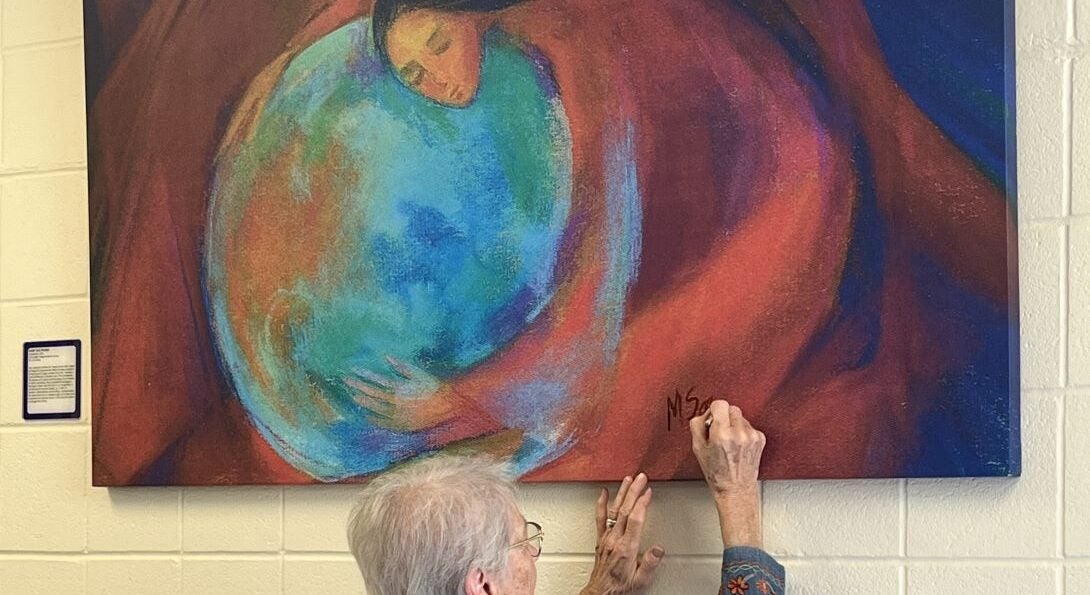 Woman signs a painting mounted to a wall of a woman embracing the earth