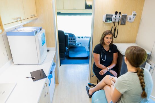 Woman with a stethoscope around her neck is sitting while speaking with a young girl whose back is to the camera.