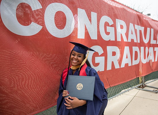 Student in cap and gown holds diploma in front of banner readying Congratulations, Graduates