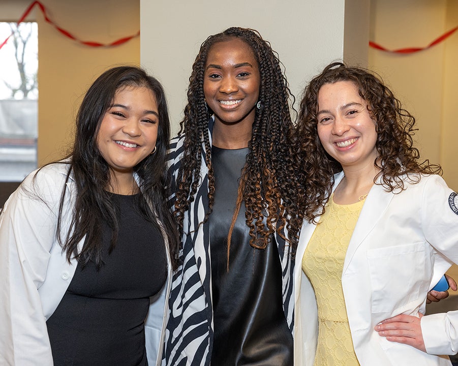 Three college-age women wearing white medical coats pose and smile at the camera