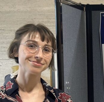 Adult student with glasses and floral blouse standing in front of poster. 