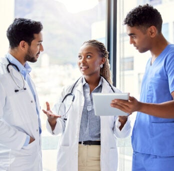 Man in white coat with stethoscope stands listening to woman in white coat with stethoscope and man in scrubs 