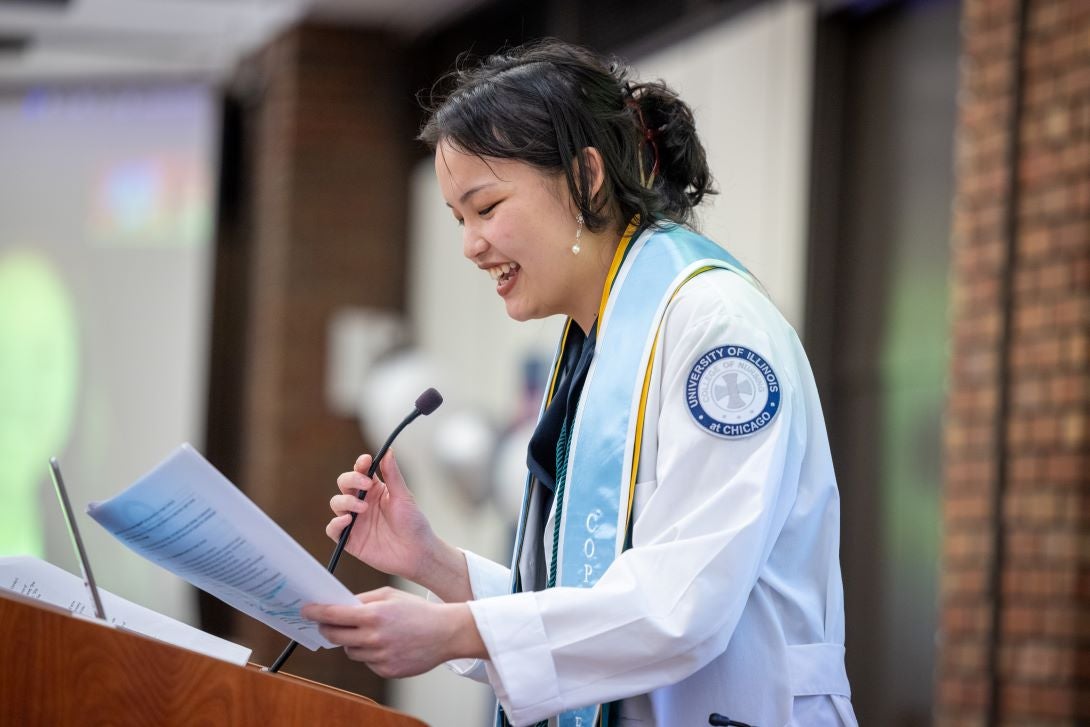 Woman with dark hair pulled back stands at a podium, holding a script and a microphone, while wearing a white coat and a blue sash around her neck
