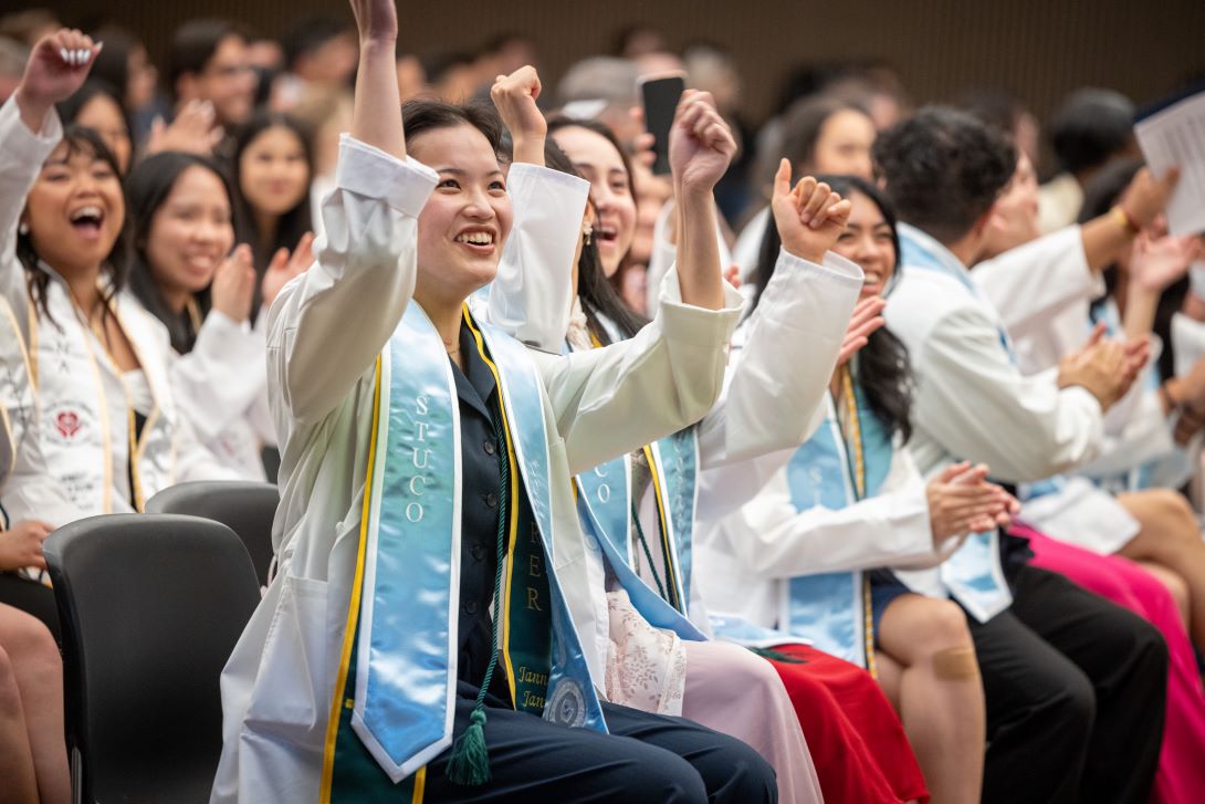 Woman in white coat with stole and cords raises up arms with smile on face