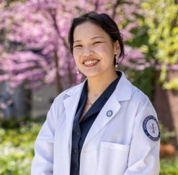 A nursing student in white coat stands smiling in front of red bud tree 