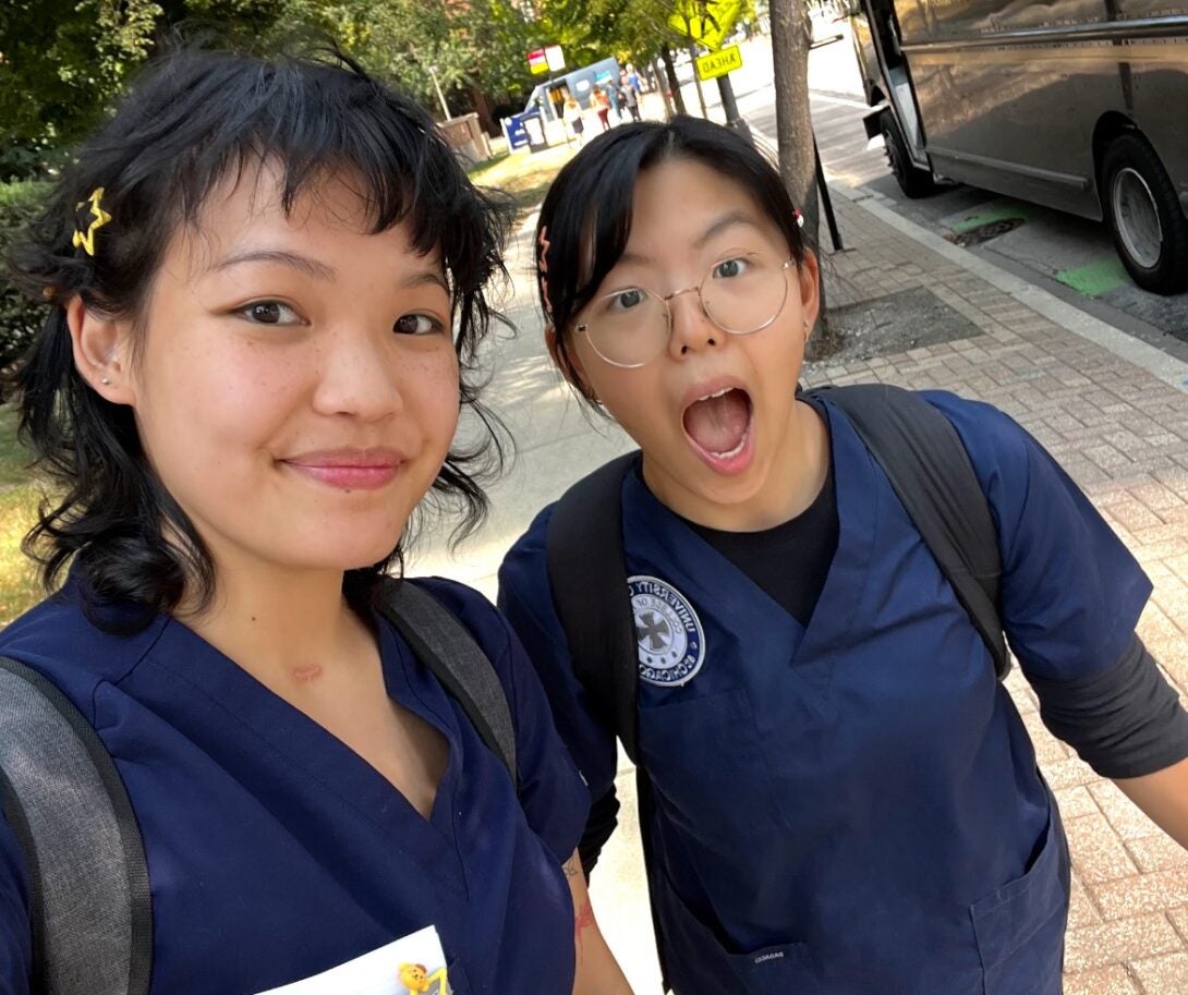 Two women in blue scrubs pose for a selfie on the sidewalk