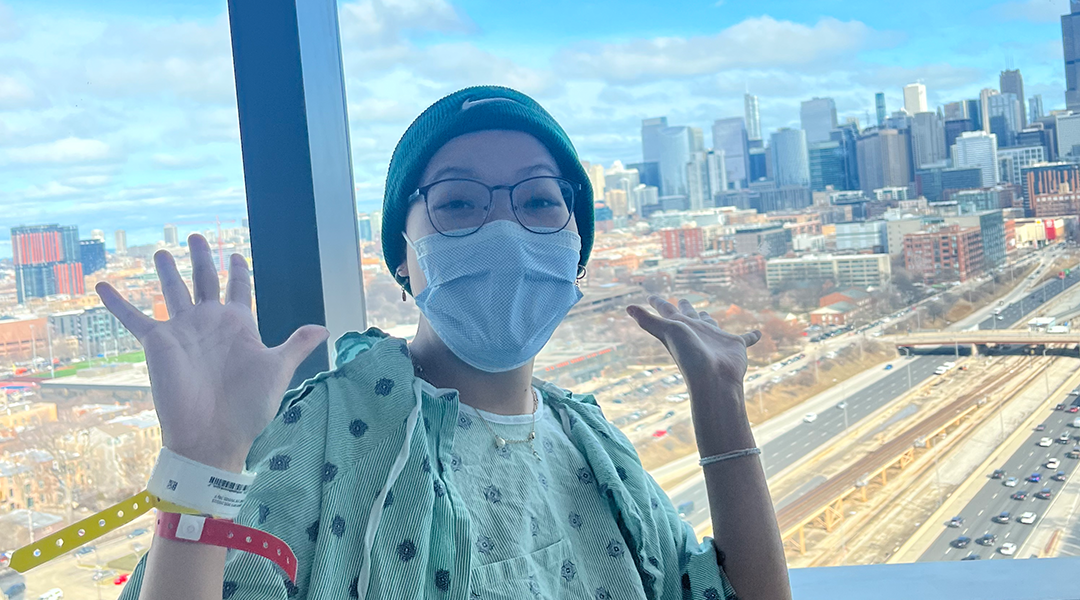 woman in hospital gown iwth mask and hat holds up hands with Chicago skyline behind her